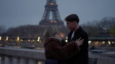 Genç bir çift, Paris 'te bir köprüde Eiffel Kulesi' nin alacakaranlıkta görülebileceği şekilde samimi bir şekilde kucaklaşır..