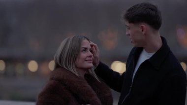 Alacakaranlıkta Paris 'te romantik bir çift erkeğin kadının saçına hafifçe dokunur sığ bir tarlanın derinliğinde arka planda bokeh ışıkları ve el kamerasıyla bir aşk ve şefkat anı yakalar..