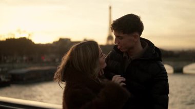 Genç bir çift Paris 'teki Seine Nehri' nin üzerindeki bir köprüde Eiffel Kulesi 'nin arka planda gün batımında görülebileceği şekilde sarılır..