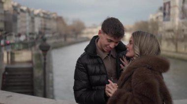 Genç bir çift Paris 'teki Seine nehri üzerindeki bir köprüde sarılır şehir binaları arka planda görünür bulutlu bir günde çift arasında romantik bir anı yakalar..