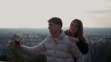 Genç bir çift arka planda Paris silueti olan bir selfie çeker kadın kolunu omzuna atarken adam telefonu tutar ve Fransa 'da altın saat boyunca romantik bir anı yakalar..