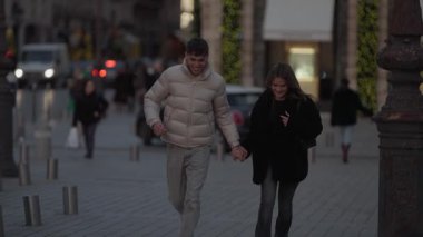 Paris sokaklarında el ele koşan genç bir çift aşıklar şehirde romantik bir akşam gezintisi yaparken gülüyor ve gülümsüyor şehir manzarası onların neşeli anlarına zemin hazırlıyor..