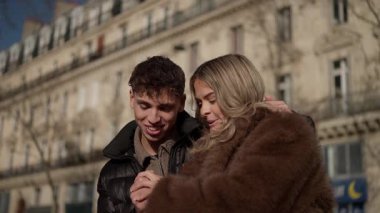 Paris 'te genç bir çift akıllı telefon ekranına bakıyor. El kameraları, güneşli bir günde Paris' teki bir binanın önünde birbirine aşık bir kadın ve erkeğin kucaklaşmalarını ve gülümsemelerini gösteriyor..