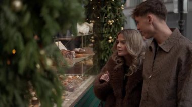 Paris 'te bir çift Noel için dekore edilmiş bir dükkanın vitrinindeki pastalara bakar kadın pastayı gösterir Fransa' da tatil alışverişini ve aşkı görüntüler..