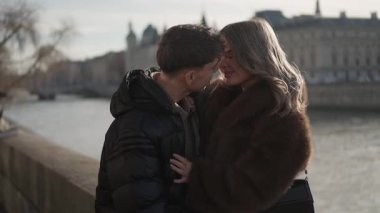 Genç bir çift Paris 'teki Seine Nehri' nin kıyısında sıkıca sarılıyor. Sahne, şehrin mimarisinin arka planda görünür olduğu romantik bir anı yakalıyor. Aşklarını ve bağlantılarını vurguluyor..