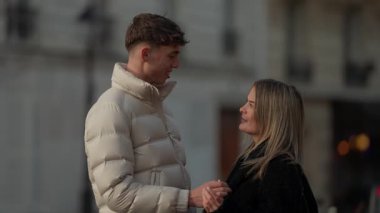 Paris 'te genç bir çift el ele tutuşup konuşuyorlar. Kadın ve erkek gülümsüyor ve birbirlerine aşık görünüyorlar. Şehir ortamında doğal ışıklandırmayla çekilmiş bir fotoğraf..