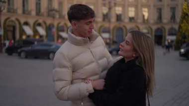 Genç bir çift Paris 'te halka açık bir meydanda el ele tutuşuyor ve kadın ve erkek birbirlerine sevgiyle bakıyor arka planda Avrupa mimarisi ve şenlikli bir atmosfer var..
