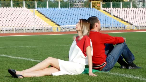 Молодой человек и красивая девушка сидит в центре футбольного поля на стадионе — стоковое видео