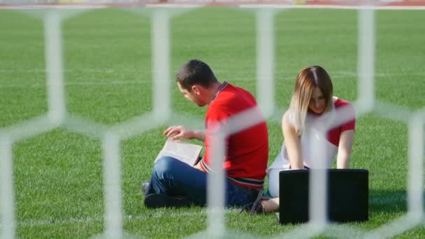 Mladý muž s knihou - hezká holka, pracují na notebooku na fotbalovém hřišti — Stock video