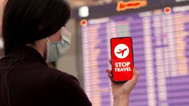 Yaklaş, kadın, koruyucu maskeli, uçak ikonlu akıllı telefon, dur işareti, ekranda, kalkış terminalinde, havaalanında uçuş bilgi panosunun önünde.
