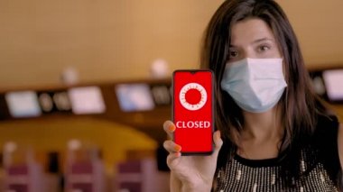 Kadın, koruyucu maskeli, elinde COVID-19 simgesi olan akıllı bir telefon tutuyor, mesaj - kapalı, ekranda, havaalanındaki boş kayıt gişelerinin önünde.