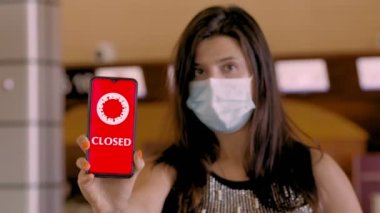 Kadın, koruyucu maskeli, elinde COVID-19 simgesi olan akıllı bir telefon tutuyor, mesaj - kapalı, ekranda, havaalanındaki boş kayıt gişelerinin önünde.