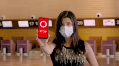 Kadın, koruyucu maskeli, elinde COVID-19 simgesi olan bir akıllı telefon tutuyor, mesaj yazıyor - ekranda, havaalanındaki boş kayıt gişelerinin önünde,.
