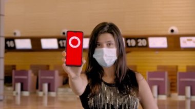 Kadın, koruyucu maskeli, elinde COVID-19 simgesi, mesajı olan akıllı telefonu var. Seyahat yasağı, ekranda, havaalanındaki boş kayıt gişelerinin önünde..