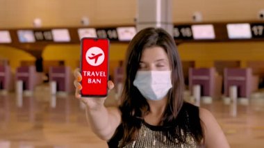 Kadın, koruyucu maskeli, elinde COVID-19 simgesi, mesajı olan akıllı telefonu var. Seyahat yasağı, ekranda, havaalanındaki boş kayıt gişelerinin önünde..