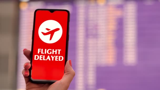 Closeup of hand holding smartphone with airplane icon, flight delayed sign, on the screen, in front of flight information board in departures terminal, at the airport. — Stock Video