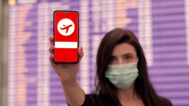 Close up, woman, in protective mask, holds smartphone with airplane icon, check in sign, on screen, in front of flight information board in departures terminal, at airport. — Stock Video