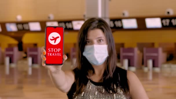 Mujer, en máscara protectora, sostiene el teléfono inteligente con el icono COVID-19, texto - detener el viaje, en la pantalla, frente a los mostradores de facturación vacíos en el aeropuerto. — Vídeo de stock