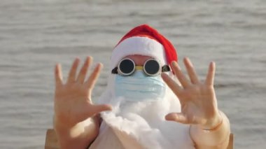 Koruyucu maskeli komik Noel Baba. Noel Baba, güneş gözlükleriyle, el sallayarak, deniz kenarında, dinleniyor. Coronavirus salgını sırasında Noel Baba tatili ya da deniz kıyısında tecrit..