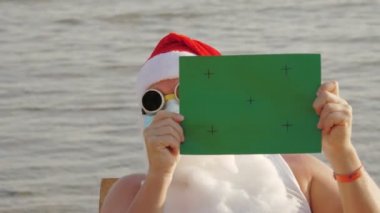 Koruyucu maskeli komik Noel Baba. Noel Baba, güneş gözlüklü, elinde yeşil bir işaret var, denizin arka planında. Coronavirus salgını sırasında Noel Baba tatili ya da deniz kıyısında tecrit..