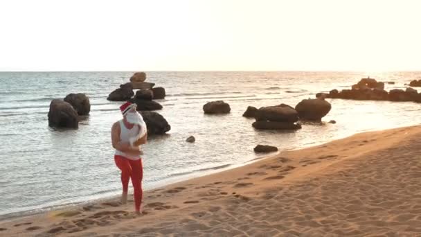 Lustig tomte. Tomten vid havet, på sommarsemester. Tomten dansar, hoppar på stranden, vid soluppgången. Tomten klor i en t-shirt och röda leggings vid stranden. — Stockvideo