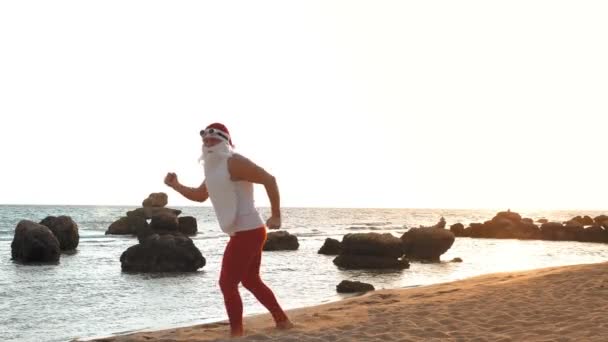 Santa Claus graciosa. Santa Claus haciendo ejercicios matutinos. Santa Claus bailando en la playa, junto al mar, al amanecer. santa claus está en camiseta y polainas rojas, en vacaciones de verano en la orilla del mar. — Vídeos de Stock