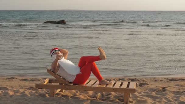 Funny santa claus sunbathes. santa lies on wooden lounger on beach by the sea and drinks a delicious cocktail. santa claus is on summer vacation, at the seashore. — Stock Video