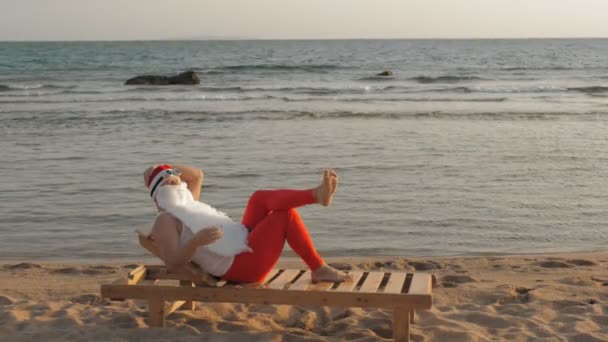 Lustige Weihnachtsmänner sonnen sich. Der Weihnachtsmann liegt auf einer Holzliege am Strand am Meer und trinkt einen köstlichen Cocktail. Weihnachtsmann ist im Sommerurlaub, am Meer. — Stockvideo