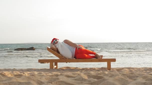 面白いサンタクラスの日光浴。サングラスのサンタは海のそばのビーチにある木製のラウンジャーに乗っています。彼は眠っていてリラックスしている。サンタ・クラスは夏休みで海岸で — ストック動画
