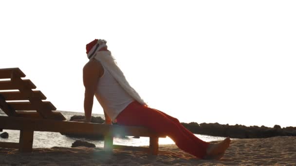 Забавные солнечные ванны Санта Клауса. Santa lies on wooden lounger on beach by the sea, relaxing, at sunrise. Санта Клаус на летних каникулах, на берегу моря. — стоковое видео