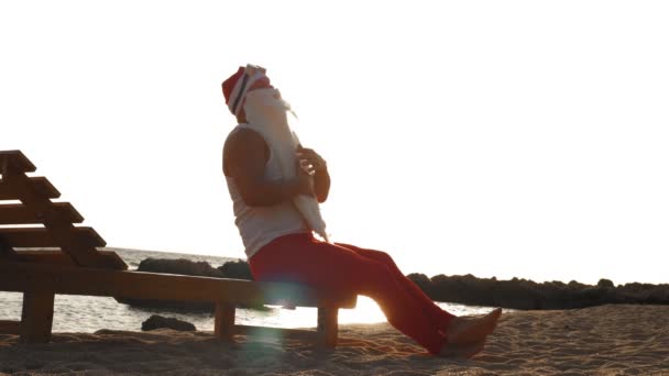 Santa claus sunbathes lucu. santa duduk di lounger kayu di pantai oleh laut, bersantai, membelai jenggot putihnya, saat matahari terbit. Santa Claus sedang berlibur musim panas, di pantai. — Stok Video