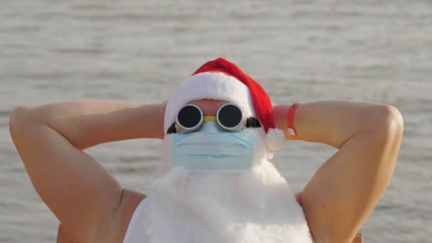 Śmieszny Święty Mikołaj w masce ochronnej. Mikołaj, w okularach przeciwsłonecznych, opalanie się na plaży nad morzem, relaks. święty Mikołaj wakacje podczas pandemii lub zamknięcia koronawirusu, na wybrzeżu. — Wideo stockowe