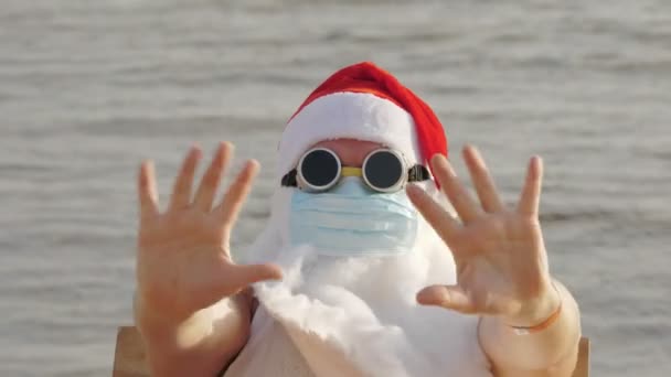 Legrační Santa Claus, v ochranné masce. Santa, ve slunečních brýlích, mává rukama, na pláži u moře, odpočívá. Santa Claus dovolená během coronavirus pandemie nebo uzamčení, na pobřeží. — Stock video
