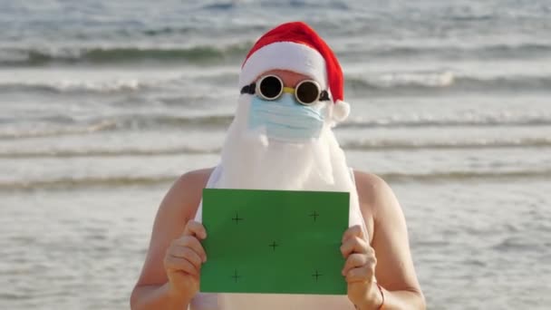 面白いサンタ・クラス保護マスクだ。サンタはサングラスをかけ、海を背景に緑の看板を掲げています。海岸でのコロナウイルスのパンデミックやロックダウン時のサンタ・クラスの休暇. — ストック動画