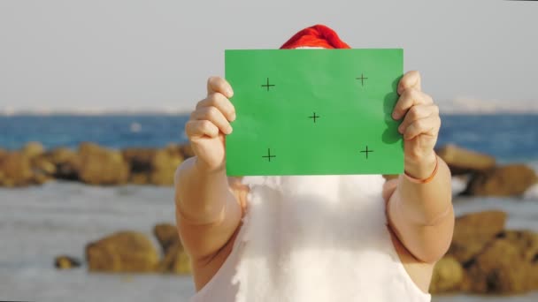 Funny santa claus, in a protective mask. Santa, in sunglasses, holding a green sign, on backdrop of the sea. santa claus vacation during coronavirus pandemic or lockdown, at the seashore. — Stock Video