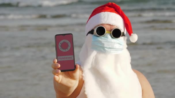 Śmieszny Święty Mikołaj w masce ochronnej. Mikołaj w okularach przeciwsłonecznych trzyma smartfona z ikoną COVID-19, tekst - pandemię, na ekranie, na plaży nad morzem. Święty Mikołaj ostrzega przed zagrożeniem ze strony koronawirusów — Wideo stockowe