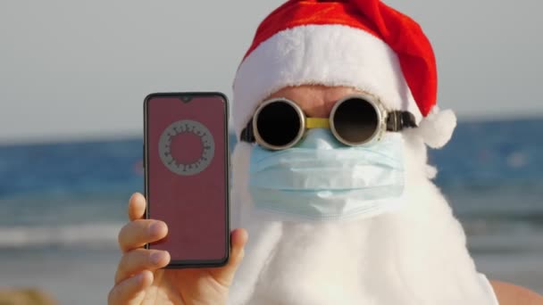Engraçado Papai Noel, com uma máscara protetora. Santa, em óculos de sol, segura smartphone com ícone COVID-19, texto - quarentena, na tela, na praia à beira-mar. Papai Noel adverte sobre o perigo de coronavírus — Vídeo de Stock