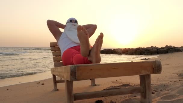 Śmieszny Mikołaj się opala. Mikołaj, w okularach przeciwsłonecznych, leży na drewnianym leżaku na plaży nad morzem, relaksujący. Święty Mikołaj jest na wakacjach, nad morzem. — Wideo stockowe