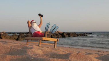 Noel Baba güneşleniyor. Komik Noel Baba, güneş gözlüğü ve palet takmış, deniz kıyısında uzanmış, elinde küçük bir müzikle müzik dinliyor, dinleniyor. Noel Baba Yazı