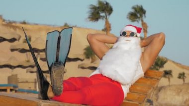 Noel Baba yaz tatili. Noel Baba güneşleniyor. Komik Noel Baba, güneş gözlükleriyle ve paletleriyle oturuyor, dinleniyor, deniz kenarında, palmiye ağaçlarına yaslanıyor..