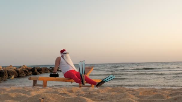 Śmieszny Mikołaj się opala. Mikołaj w okularach przeciwsłonecznych i płetwach siedzi na leżaku, na plaży nad morzem i słucha muzyki, relaksuje się. Święty Mikołaj letnie wakacje, nad morzem. — Wideo stockowe