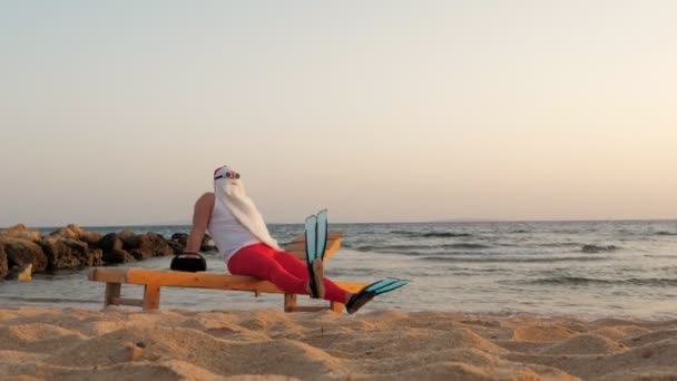 Αστεία sunbathes του Άη Βασίλη. Ο Άγιος Βασίλης, με γυαλιά ηλίου και βατραχοπέδιλα, κάθεται στο lounger, στην παραλία δίπλα στη θάλασσα και ακούει μουσική, χαλαρώνοντας. Άγιος Βασίλης καλοκαιρινές διακοπές, στην παραλία. — Αρχείο Βίντεο