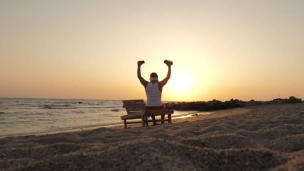 Der Weihnachtsmann tanzt. Lustiger Weihnachtsmann, der mit einem drahtlosen Bluetooth-Lautsprecher in der Hand vor Sonnenuntergang oder Sonnenaufgang tanzt. Rückseite. Entspannen Sie sich am Strand am Meer — Stockvideo