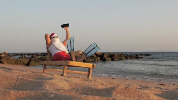 Santa Claus sunbathes. Funny Santa Claus, in sunglasses and flippers, is sitting on lounger, on beach by the sea and listening to music with small musical speaker in hand, relaxing. santa claus summer — Stock Video