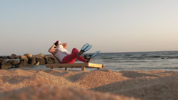 Санта Клаус загорает. Забавный Санта Клаус, в солнечных очках и ластах, сидит на шезлонге, на пляже у моря и слушает музыку с небольшим музыкальным динамиком в руке, расслабляясь. Санта-Клаус летом — стоковое видео