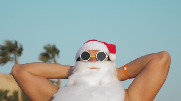 Der Weihnachtsmann im Sommerurlaub. Porträt. Der Nikolaus sonnt sich. Lustiger Weihnachtsmann mit Sonnenbrille sitzt entspannt am Strand am Meer vor Palmen-Kulisse. — Stockvideo