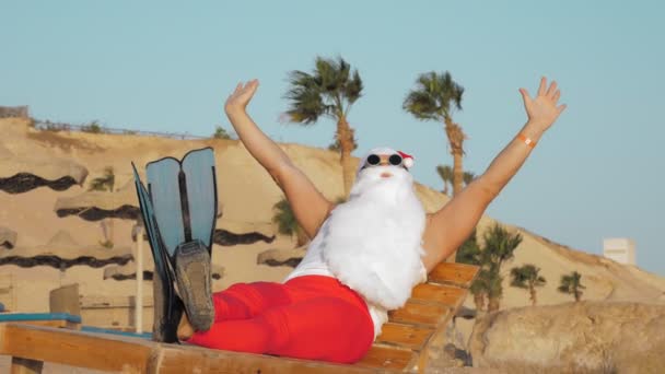 Vacaciones de verano de Papá Noel. Santa Claus toma el sol. Funny Santa, en gafas de sol y aletas, está sentado, relajándose en la tumbona, en la playa junto al mar, contra palmeras telón de fondo. — Vídeos de Stock