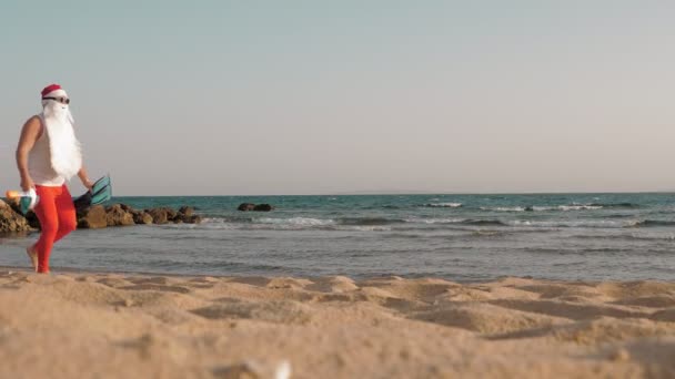 Der Weihnachtsmann im Sommerurlaub. Der Weihnachtsmann hat Spaß. Der lustige Weihnachtsmann mit Sonnenbrille, Schwimmflossen und Schnorchelmaske läuft am Strand entlang. Er hat es eilig zu schwimmen, zu schnorcheln. — Stockvideo