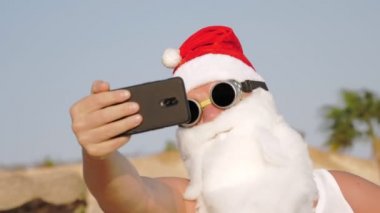 Noel Baba selfie çeker. Noel Baba yaz tatili. Noel Baba blogcusu. Güneş gözlüklü komik Noel Baba, sahilde palmiye ağaçlarına karşı selfie videosu çekiyor..