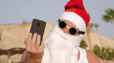 Noel Baba selfie çeker. Noel Baba yaz tatili. Noel Baba blogcusu. Güneş gözlüklü komik Noel Baba, sahilde palmiye ağaçlarına karşı selfie videosu çekiyor..